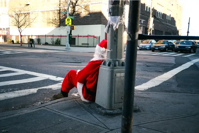 A person dressed as Santa Claus, sitting on a street corner with his back turned to the camera.